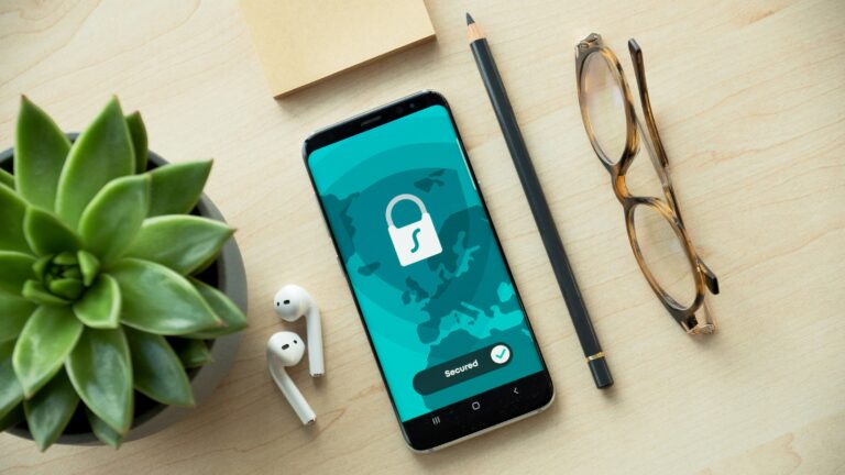 Smartphone mit Schloss-Symbol auf einem Schreibtisch neben Brille, Stift und Pflanze.